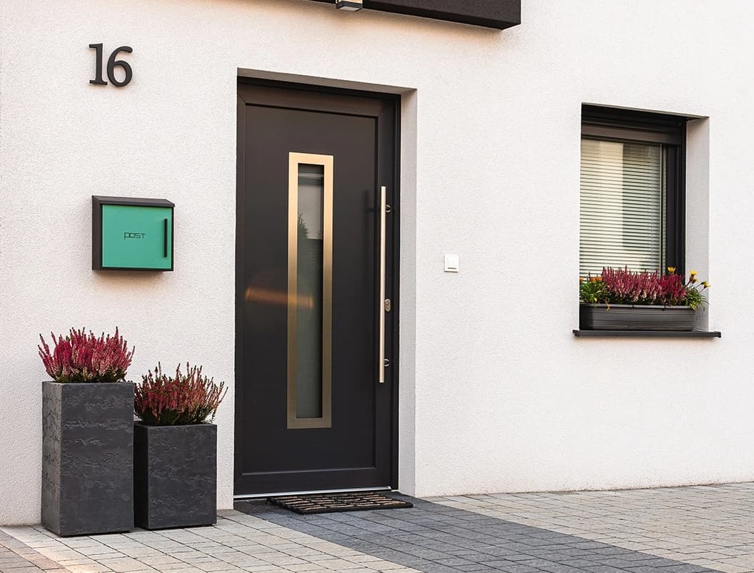 Ciemne drzwi wejściowe PVC Modern Line z numerem 16 na ścianie, otoczone ozdobnymi donicami