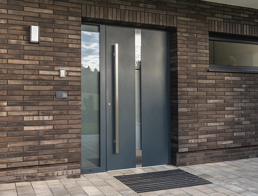 Nowoczesne aluminiowe drzwi wejściowe Premium Line z przeszkleniami, wkomponowane w ciemną ceglaną elewację.